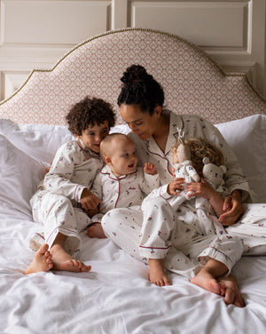 A family of four wearing matching white patterned pajamas sits together on a bed with white linens and a patterned upholstered headboard. The woman holds a doll, and the children look toward the camera.