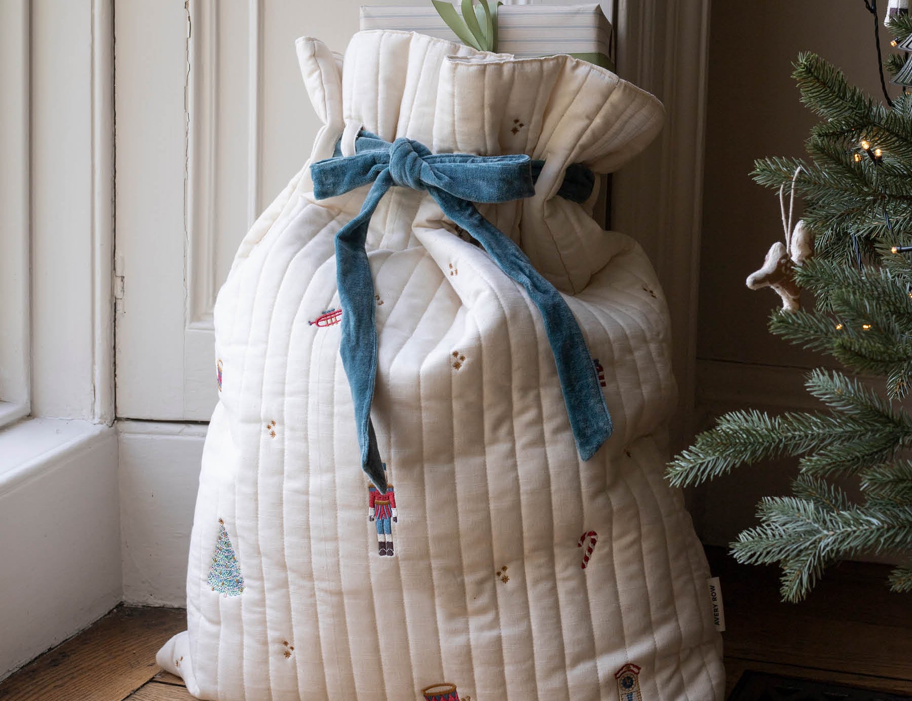 A large quilted Christmas stocking bag in cream with a blue velvet bow and nutcracker print, standing on a wooden floor next to a Christmas tree.