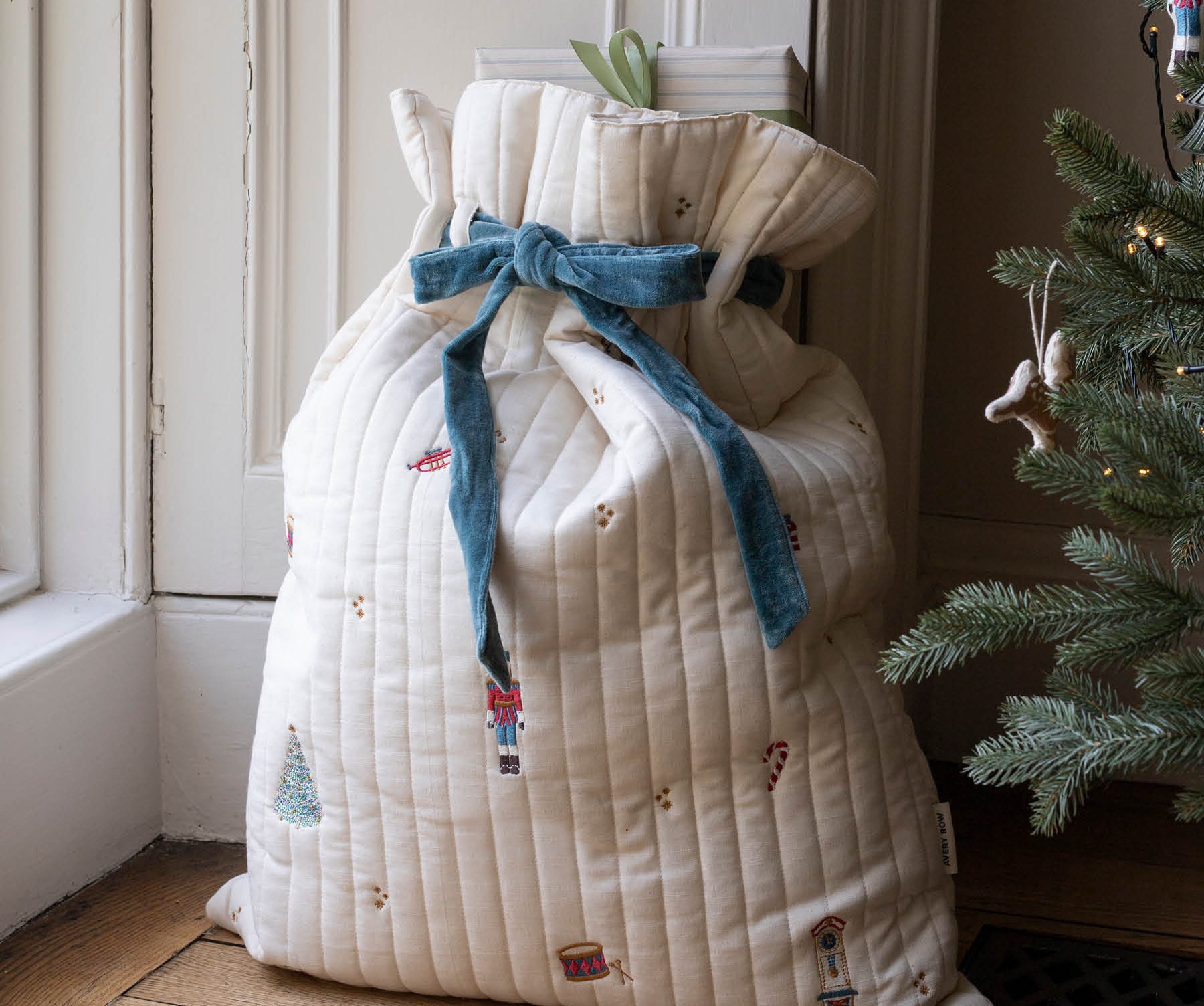 A large quilted Christmas stocking bag in cream with a blue velvet bow and nutcracker print, standing on a wooden floor next to a Christmas tree.