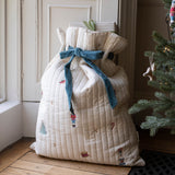 A large quilted Christmas stocking bag in cream with a blue velvet bow and nutcracker print, standing on a wooden floor next to a Christmas tree.