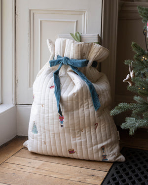 A large quilted Christmas stocking bag in cream with a blue velvet bow and nutcracker print, standing on a wooden floor next to a Christmas tree.