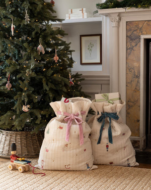 Two quilted cream fabric gift bags with festive prints and ribbon bows, placed on a woven rug in front of a decorated Christmas tree and fireplace.