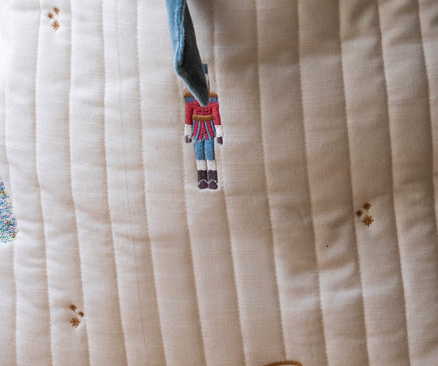 Close-up of a quilted cream fabric cushion with vertical channel stitching, embroidered nutcracker and drum motifs, and a blue drawstring.