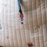 Close-up of a quilted cream fabric cushion with vertical channel stitching, embroidered nutcracker and drum motifs, and a blue drawstring.