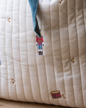 Close-up of a quilted cream fabric cushion with vertical channel stitching, embroidered nutcracker and drum motifs, and a blue drawstring.