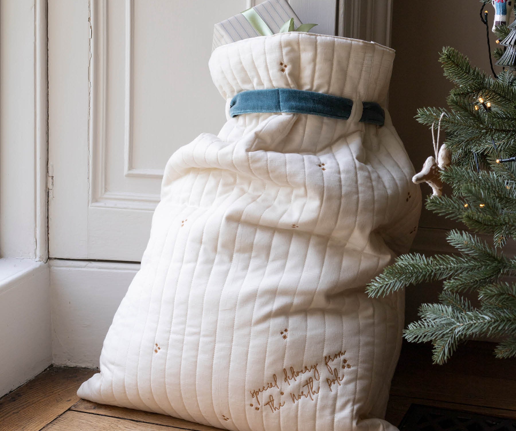 A large quilted cream Christmas stocking or gift sack with embroidered details and a blue ribbon, standing on a wooden floor next to a Christmas tree.
