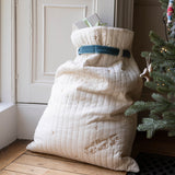 A large quilted cream Christmas stocking or gift sack with embroidered details and a blue ribbon, standing on a wooden floor next to a Christmas tree.
