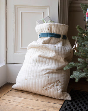 A large quilted cream Christmas stocking or gift sack with embroidered details and a blue ribbon, standing on a wooden floor next to a Christmas tree.