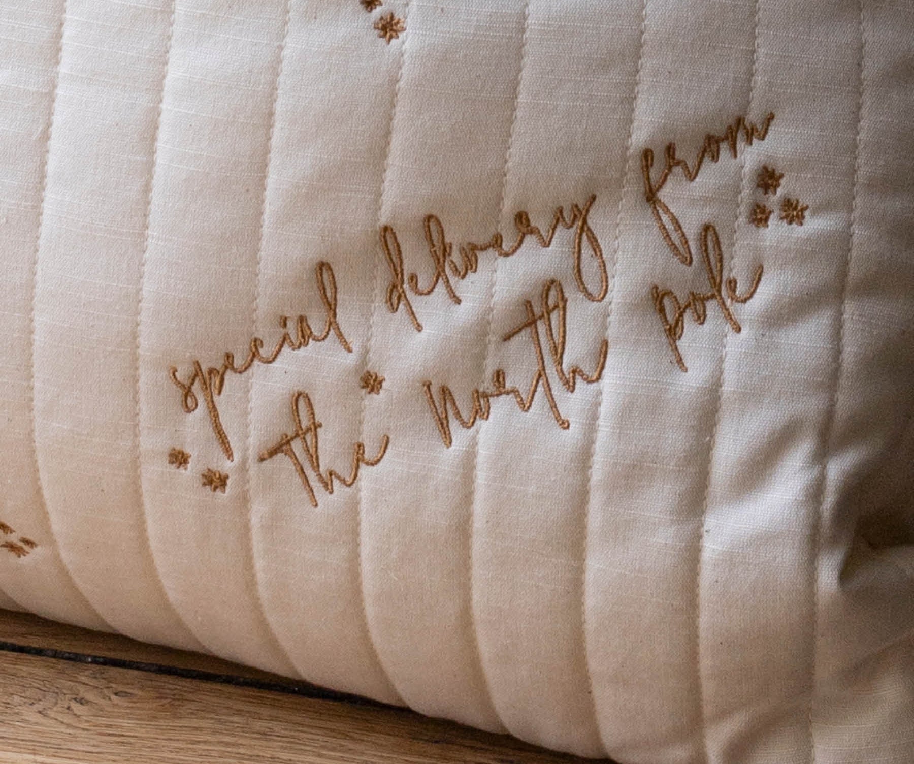 A close-up of a cream-colored quilted pillow with brown embroidered text reading 'special delivery from The North Pole' and small star motifs, resting on a wooden surface.