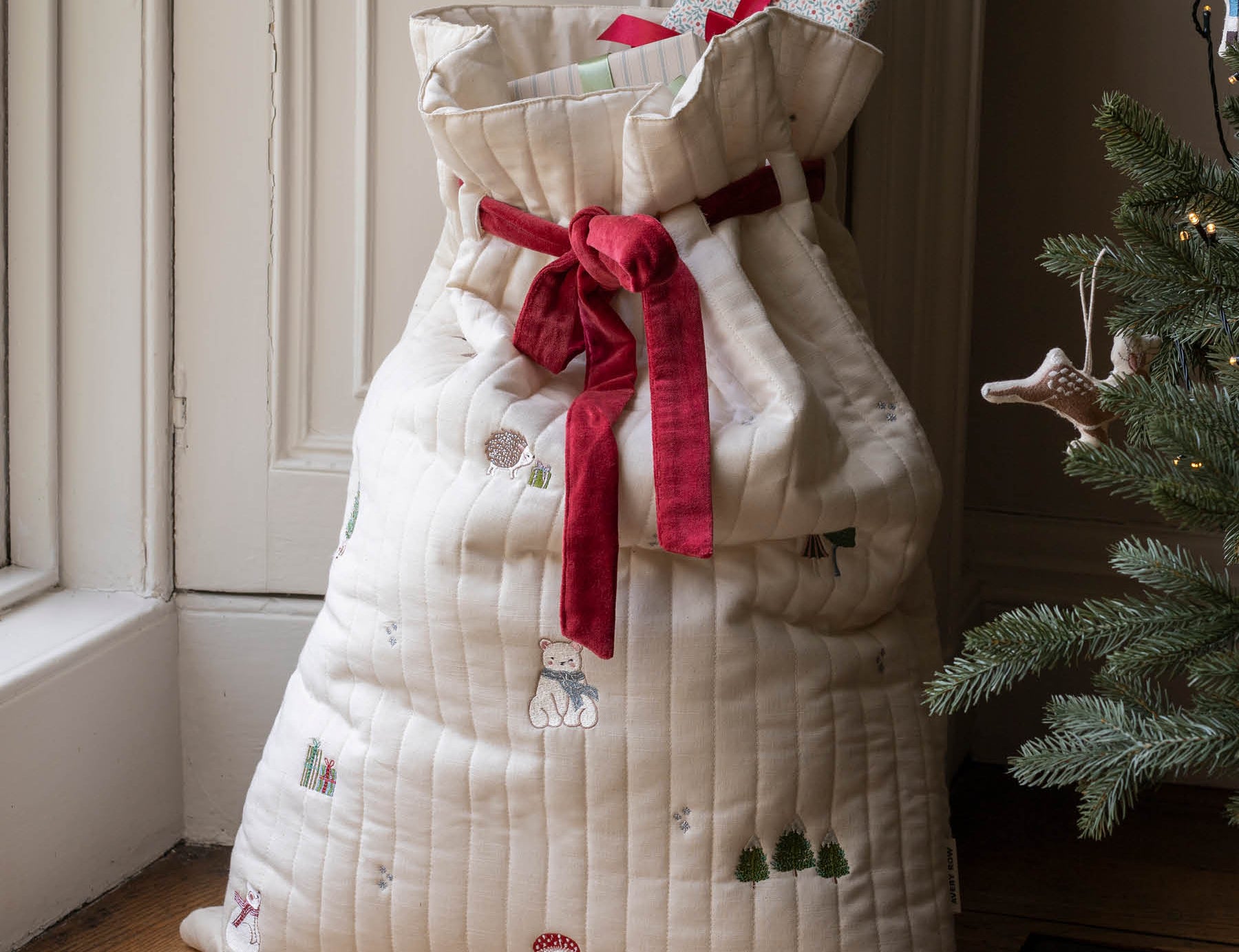 A large quilted Christmas stocking filled with wrapped presents, tied with a red ribbon, standing on a wooden floor next to a Christmas tree.