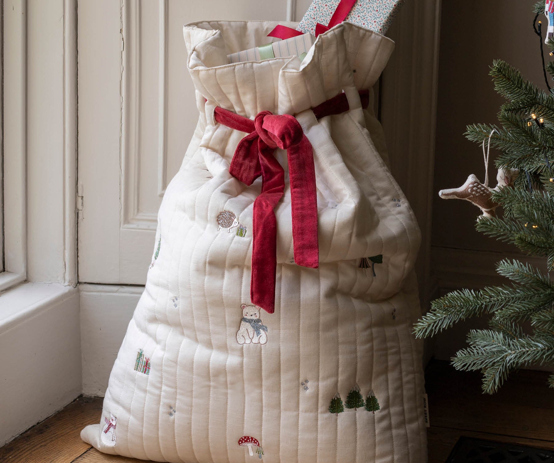 A large quilted Christmas stocking filled with wrapped presents, tied with a red ribbon, standing on a wooden floor next to a Christmas tree.