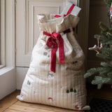 A large quilted Christmas stocking filled with wrapped presents, tied with a red ribbon, standing on a wooden floor next to a Christmas tree.