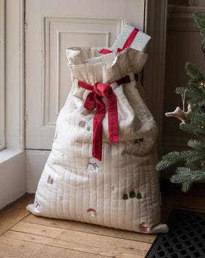 A large quilted Christmas stocking filled with wrapped presents, tied with a red ribbon, standing on a wooden floor next to a Christmas tree.