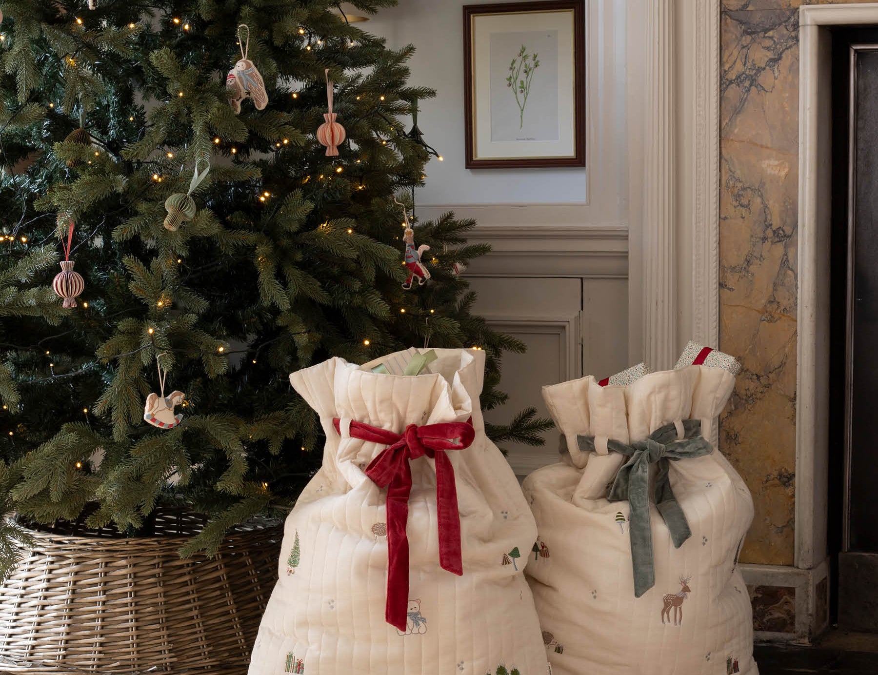 Two cream-colored quilted fabric gift bags with holiday prints and drawstring tops, one tied with a red ribbon and the other with a gray ribbon, displayed on a woven rug in front of a decorated Christmas tree and fireplace.