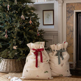 Two cream-colored quilted fabric gift bags with holiday prints and drawstring tops, one tied with a red ribbon and the other with a gray ribbon, displayed on a woven rug in front of a decorated Christmas tree and fireplace.