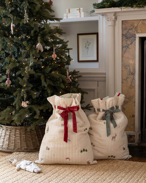 Two cream-colored quilted fabric gift bags with holiday prints and drawstring tops, one tied with a red ribbon and the other with a gray ribbon, displayed on a woven rug in front of a decorated Christmas tree and fireplace.