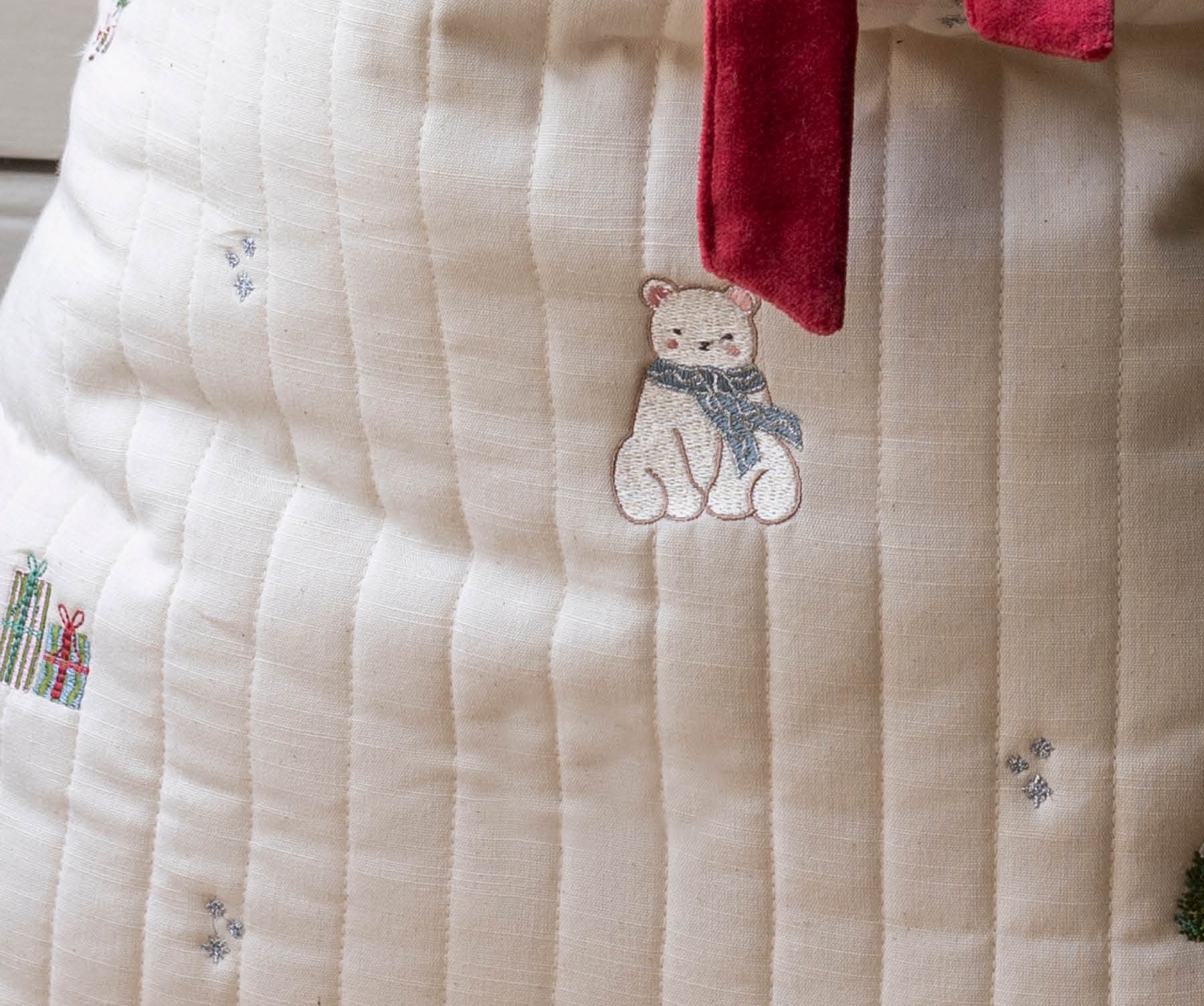 Close-up of a cream-colored quilted fabric item, likely a blanket or cushion cover, featuring embroidered details of a bear wearing a blue scarf, a red mushroom, and small gift boxes, with a red velvet tie at the top.