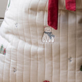 Close-up of a cream-colored quilted fabric item, likely a blanket or cushion cover, featuring embroidered details of a bear wearing a blue scarf, a red mushroom, and small gift boxes, with a red velvet tie at the top.