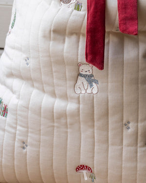Close-up of a cream-colored quilted fabric item, likely a blanket or cushion cover, featuring embroidered details of a bear wearing a blue scarf, a red mushroom, and small gift boxes, with a red velvet tie at the top.
