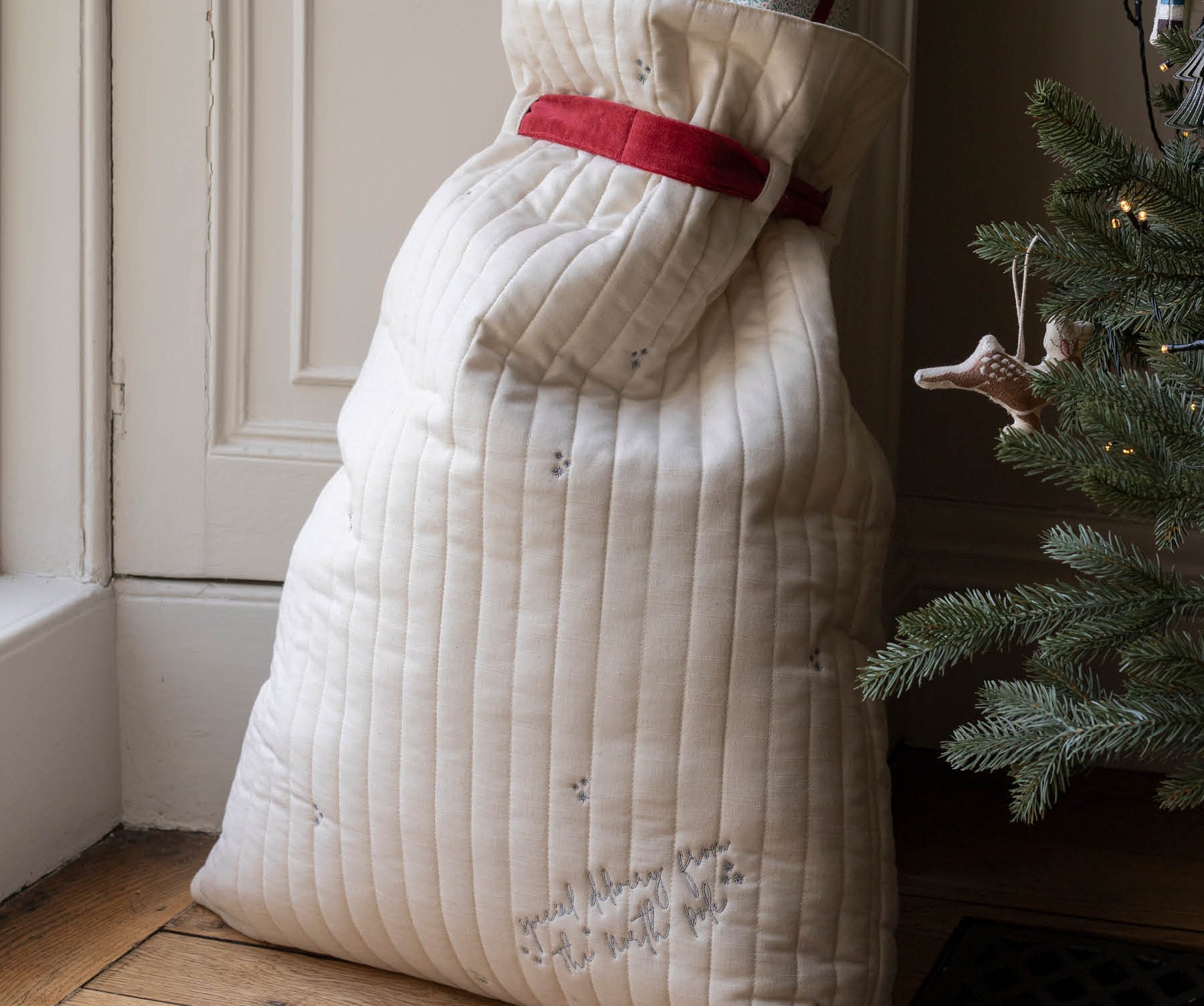 A large quilted Christmas stocking filled with gifts, standing on a wooden floor next to a Christmas tree. The stocking is cream-colored with a red waistband and subtle embroidered details.
