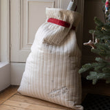A large quilted Christmas stocking filled with gifts, standing on a wooden floor next to a Christmas tree. The stocking is cream-colored with a red waistband and subtle embroidered details.