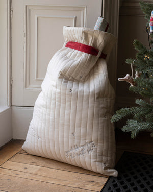 A large quilted Christmas stocking filled with gifts, standing on a wooden floor next to a Christmas tree. The stocking is cream-colored with a red waistband and subtle embroidered details.