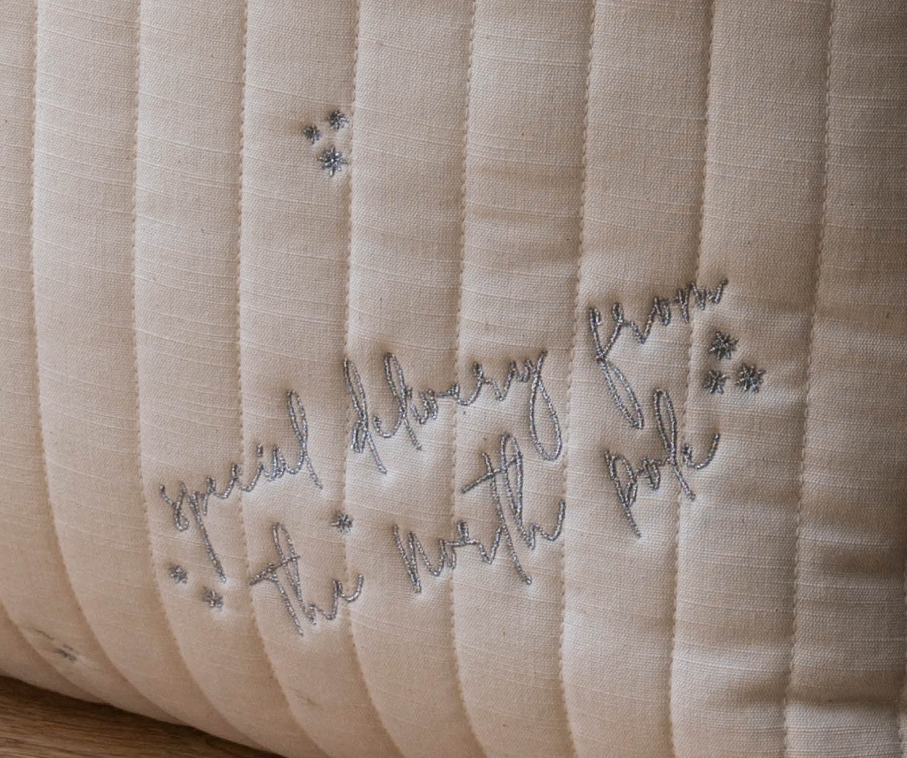 Close-up of a quilted cream-colored pillow with vertical channel stitching and embroidered gray text reading 'special delivery from the north pole' with small star accents, resting on a wooden surface.