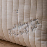 Close-up of a quilted cream-colored pillow with vertical channel stitching and embroidered gray text reading 'special delivery from the north pole' with small star accents, resting on a wooden surface.