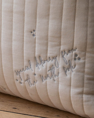 Close-up of a quilted cream-colored pillow with vertical channel stitching and embroidered gray text reading 'special delivery from the north pole' with small star accents, resting on a wooden surface.