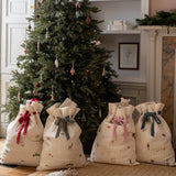 Four cream quilted Christmas stocking bags with festive prints and red, green, pink, and blue ribbons tied at the top, arranged on a woven rug in front of a decorated Christmas tree in a cozy room.