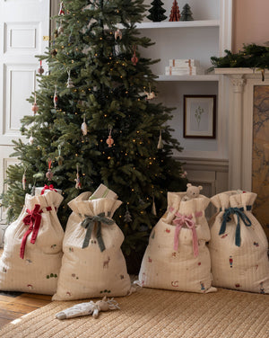 Four cream quilted Christmas stocking bags with festive prints and red, green, pink, and blue ribbons tied at the top, arranged on a woven rug in front of a decorated Christmas tree in a cozy room.
