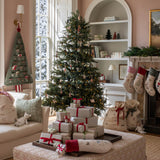 A beautifully decorated Christmas living room featuring a tall green tree with ornaments, wrapped gift boxes on a patterned ottoman, stockings hanging from a fireplace, and a cozy armchair.