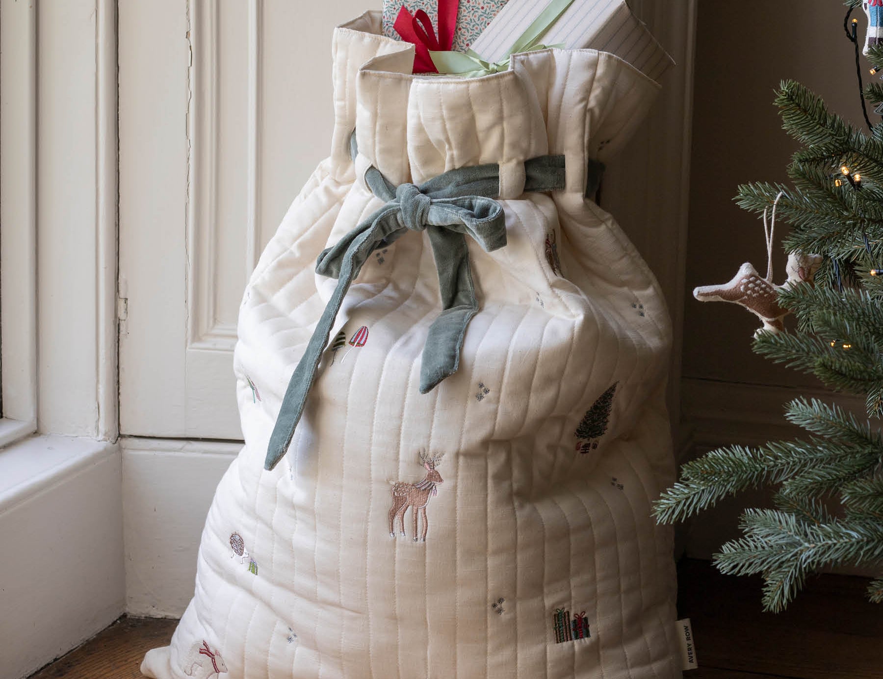 A large quilted Christmas stocking or gift bag in cream fabric with embroidered holiday motifs, tied with a sage green ribbon, standing on a wooden floor next to a Christmas tree and doorway.