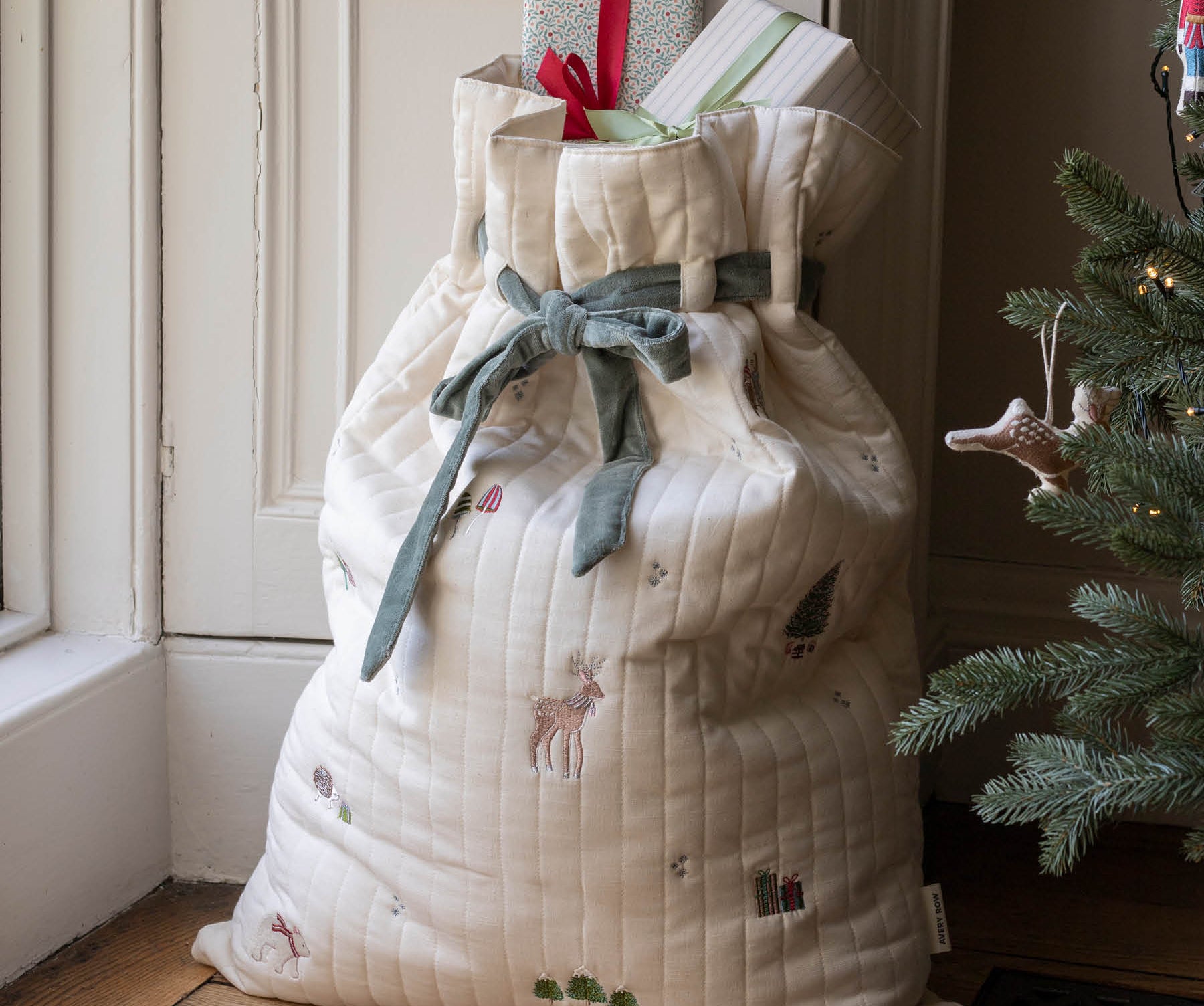 A large quilted Christmas stocking or gift bag in cream fabric with embroidered holiday motifs, tied with a sage green ribbon, standing on a wooden floor next to a Christmas tree and doorway.