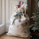 A large quilted Christmas stocking or gift bag in cream fabric with embroidered holiday motifs, tied with a sage green ribbon, standing on a wooden floor next to a Christmas tree and doorway.