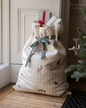A large quilted Christmas stocking or gift bag in cream fabric with embroidered holiday motifs, tied with a sage green ribbon, standing on a wooden floor next to a Christmas tree and doorway.