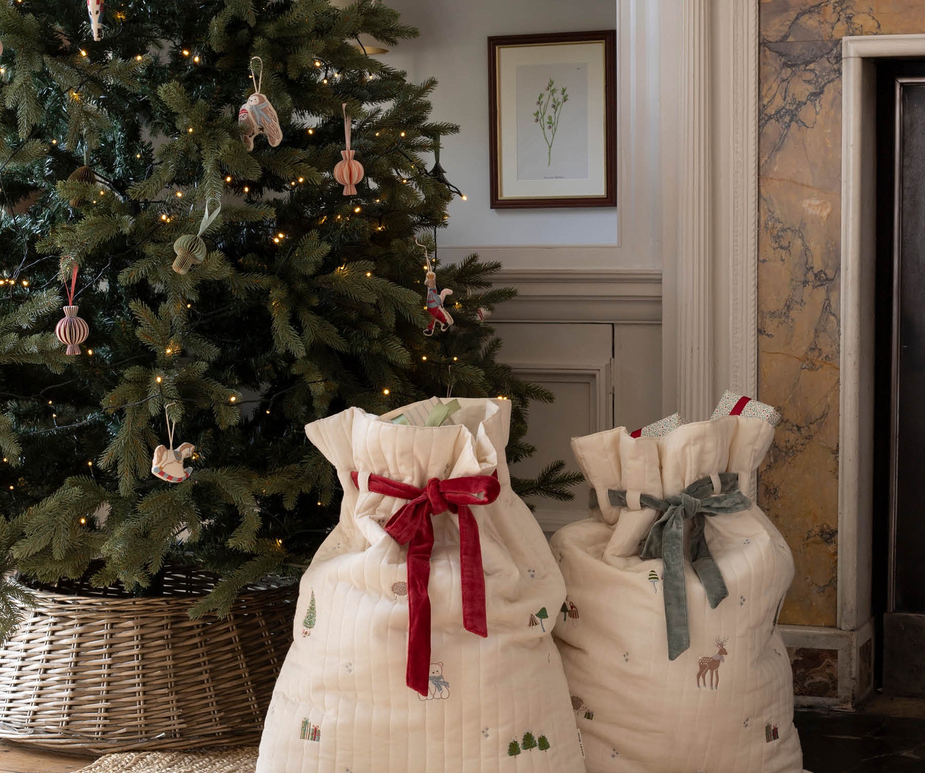 Two cream-colored quilted fabric gift bags with holiday prints and drawstring tops, one tied with a red ribbon and the other with a gray ribbon, displayed on a woven rug in front of a decorated Christmas tree and fireplace.