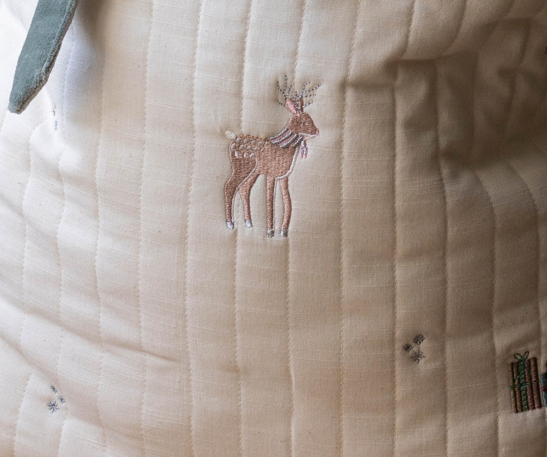 Close-up of a quilted cream fabric item, likely a baby blanket or cushion cover, featuring embroidered details of a brown deer, small green trees, and tiny blue floral motifs.