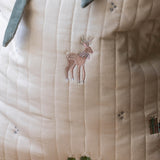 Close-up of a quilted cream fabric item, likely a baby blanket or cushion cover, featuring embroidered details of a brown deer, small green trees, and tiny blue floral motifs.
