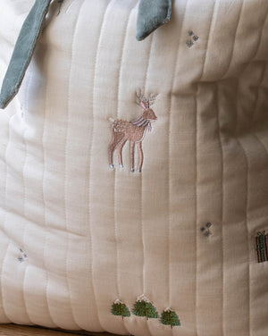 Close-up of a quilted cream fabric item, likely a baby blanket or cushion cover, featuring embroidered details of a brown deer, small green trees, and tiny blue floral motifs.