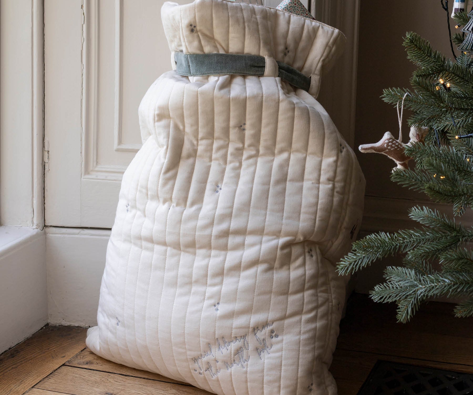 A large quilted cream-colored Christmas stocking filled with blankets or quilts, tied with a gray ribbon, standing on a wooden floor next to a Christmas tree.