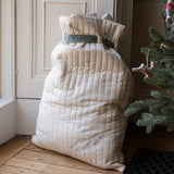 A large quilted cream-colored Christmas stocking filled with blankets or quilts, tied with a gray ribbon, standing on a wooden floor next to a Christmas tree.