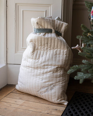 A large quilted cream-colored Christmas stocking filled with blankets or quilts, tied with a gray ribbon, standing on a wooden floor next to a Christmas tree.