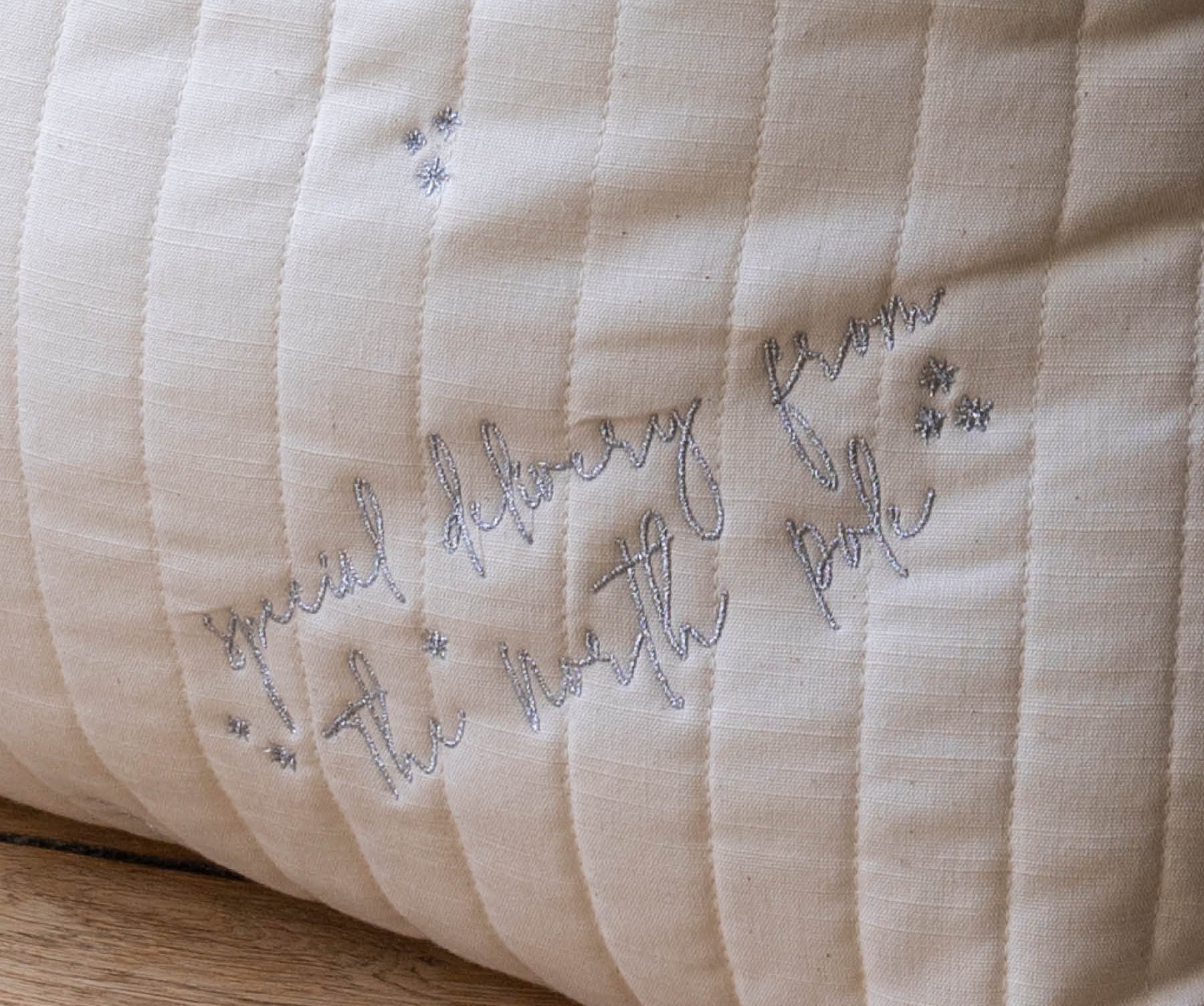 A close-up of a cream-colored quilted fabric cushion with vertical channel stitching and embroidered cursive text reading 'Special delivery from the north pole' in gray thread, resting on a wooden surface.