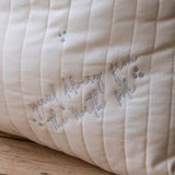 A close-up of a cream-colored quilted fabric cushion with vertical channel stitching and embroidered cursive text reading 'Special delivery from the north pole' in gray thread, resting on a wooden surface.