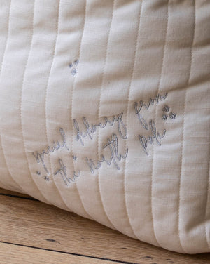 A close-up of a cream-colored quilted fabric cushion with vertical channel stitching and embroidered cursive text reading 'Special delivery from the north pole' in gray thread, resting on a wooden surface.