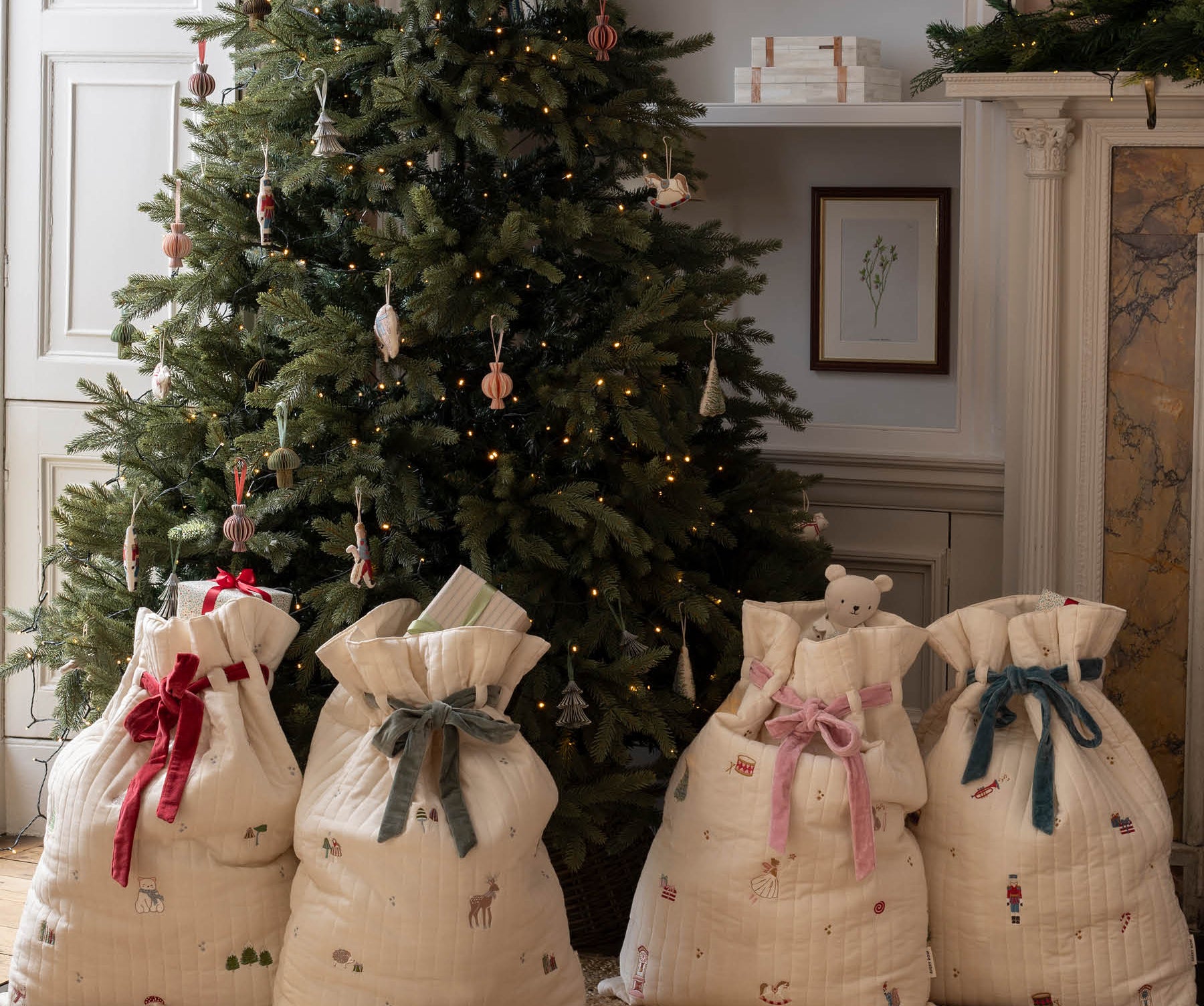 Four cream quilted Christmas stocking bags with festive prints and red, green, pink, and blue ribbons tied at the top, arranged on a woven rug in front of a decorated Christmas tree in a cozy room.