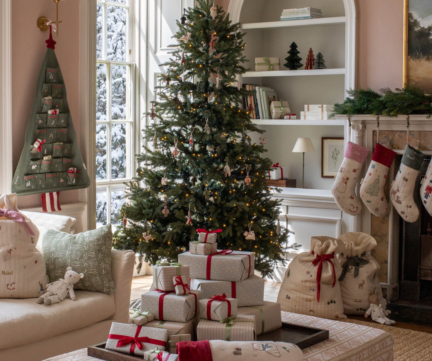 A beautifully decorated Christmas living room featuring a tall green tree with ornaments, wrapped gift boxes on a patterned ottoman, stockings hanging from a fireplace, and a cozy armchair.