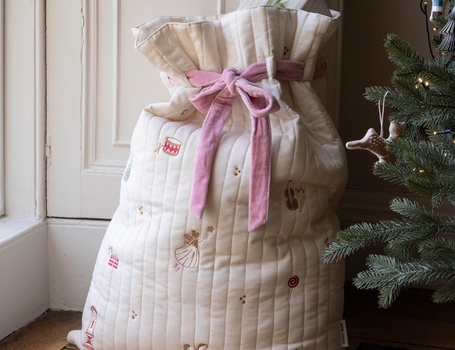 A quilted cream Christmas stocking filled with gifts, tied with a pink ribbon, standing on a wooden floor next to a decorated Christmas tree.