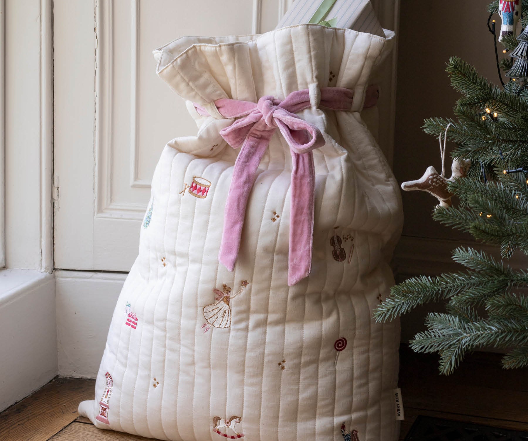 A quilted cream Christmas stocking filled with gifts, tied with a pink ribbon, standing on a wooden floor next to a decorated Christmas tree.